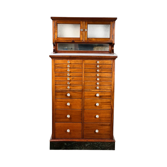 An antique mahogany multi-drawer dental cabinet by The American Cabinet Co., early 20th century, with a black base, two frosted glass upper doors, pull-out writing surface, and small drawers with round white knobs.