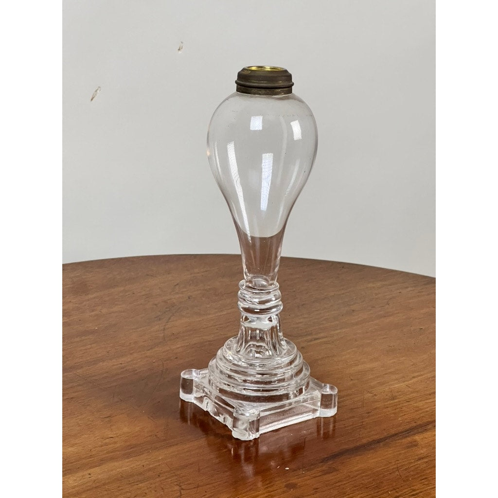 An Attributed Boston & Sandwich Pressed Glass Boudoir Fluid Lamp, Circa 1850, with a clear glass base featuring a bulbous top inspired by early American lighting, stands on an ornate square pedestal atop a wooden surface against a plain light background. This elegant piece captures the mid-19th century style characteristic of fluid lamps linked to the famed Boston & Sandwich Glass Company.
