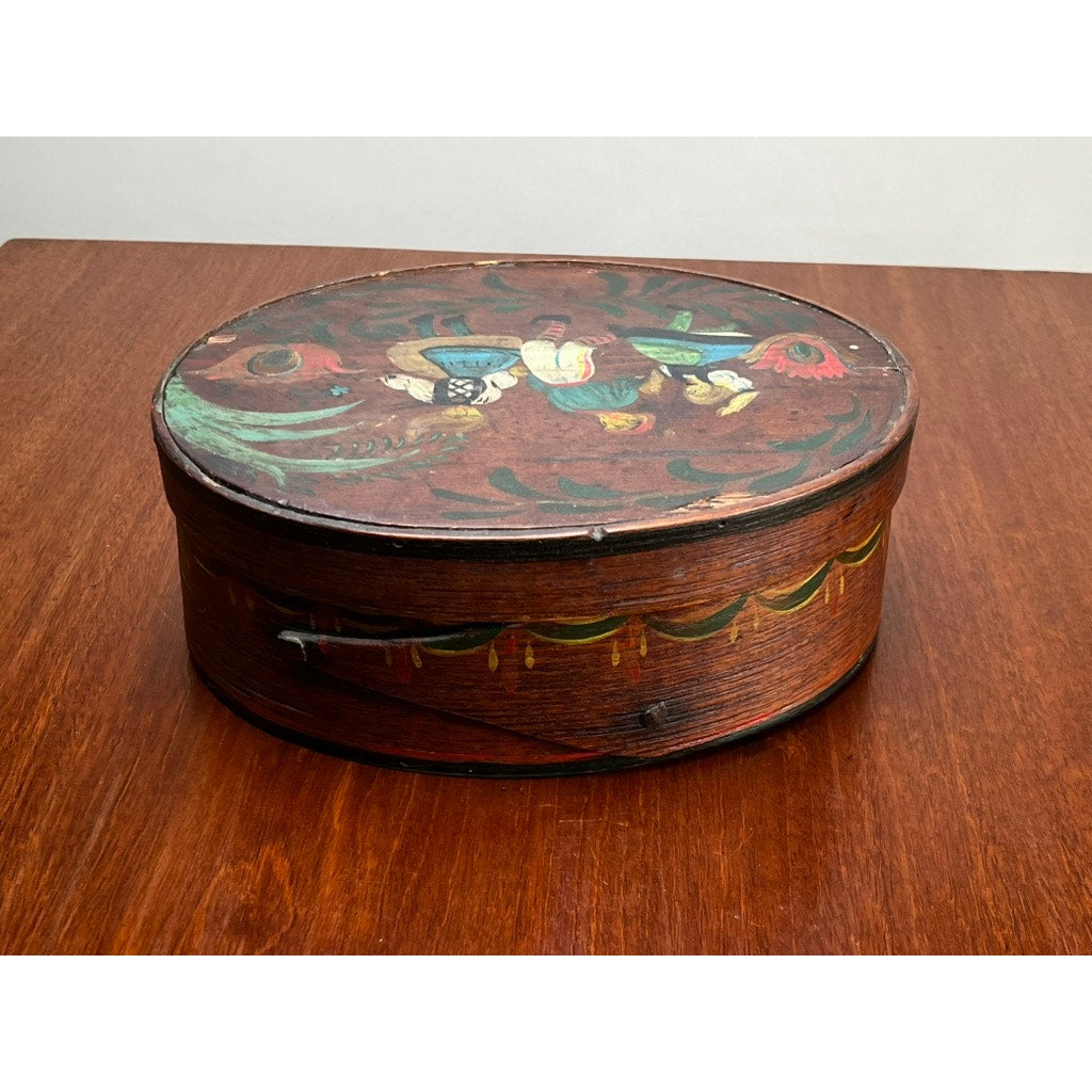 An Antique German polychrome painted bentwood "Spanschachtel" ribbon box, late 18th century, featuring floral and bird motifs on the lid with green and yellow side accents, sits atop a brown wooden surface. Brand unknown.