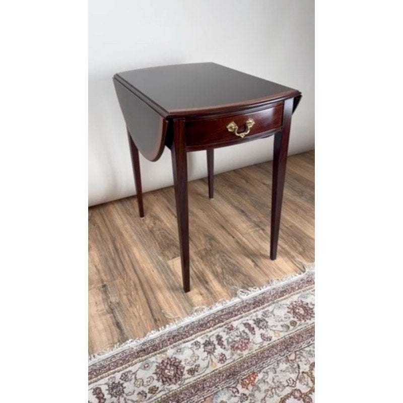 A Vintage Federal Style Edge Banded Mahogany End Table by Stickley, featuring a single brass-handled drawer, stands on a hardwood floor beside a patterned rug.