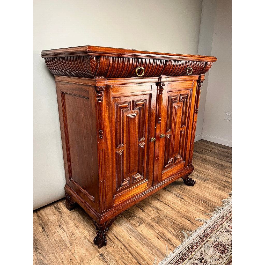 Antique English Jacobean Revival Mahogany Buffet c. 1915–1925 with ornate carvings, two paneled doors, brass handles, and claw feet stands on a wooden floor beside a patterned rug and plain white wall. Brand: None/Unknown.