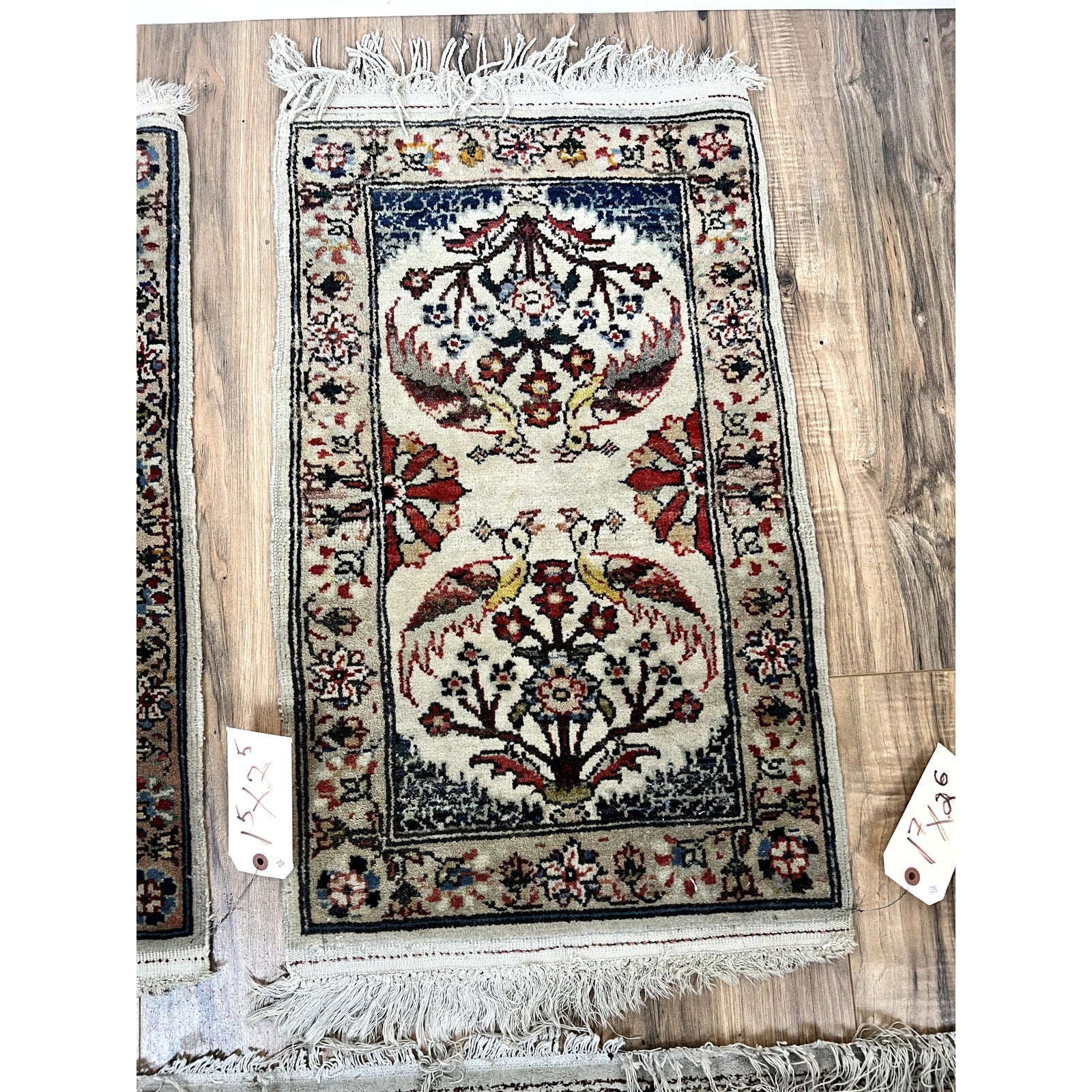 A pair of vintage hand-knotted Oriental "Tree of Life" scatter rugs (1.5 x 2.5), featuring intricate patterns and bird motifs on a cream background, with fringed edges and two price tags, is displayed on a wooden floor. Brand unknown.