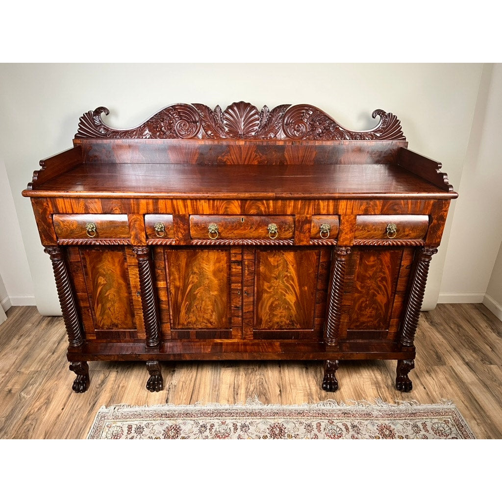 Antique American Empire Mahogany Sideboard with rosewood edge banding, likely Philadelphia c.1840, features carved detailing, four drawers with brass handles, twisted columns, and claw feet. Shown on a wooden floor with a patterned rug in front.