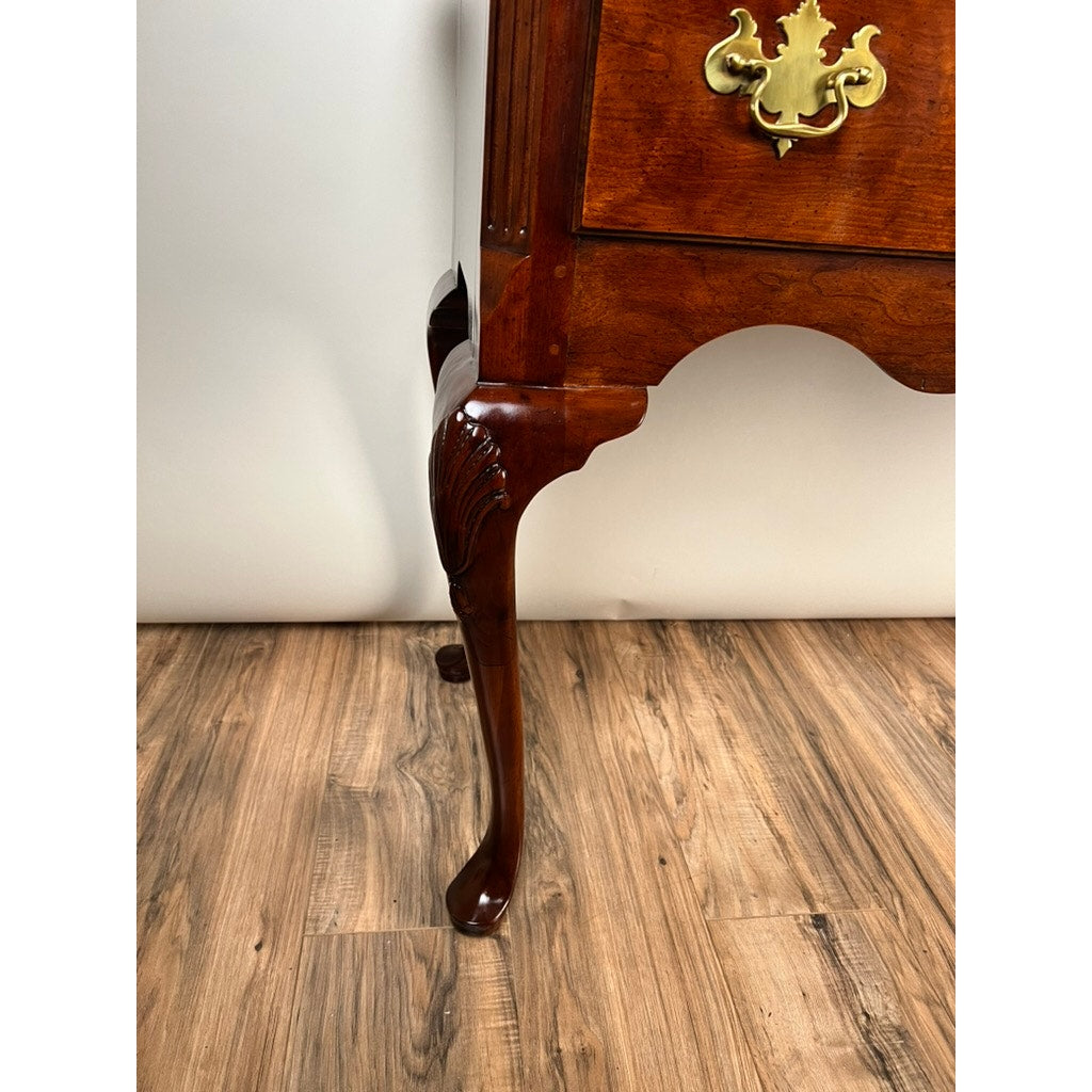 Close-up of a Vintage Solid Cherry Queen Anne Style Sideboard by Henredon, featuring a curved carved leg and ornate brass handle, placed on a wood-patterned floor against a plain light-colored wall.