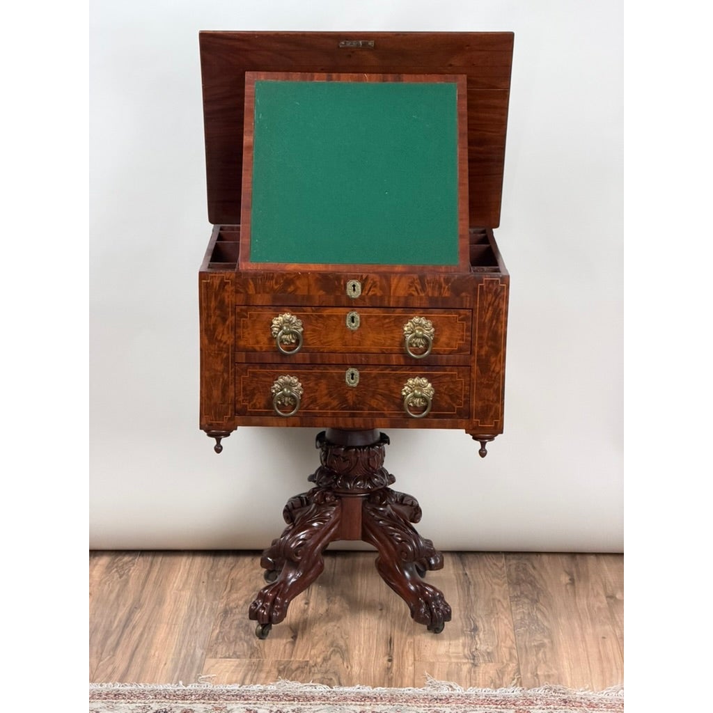 An American Classical Figured Mahogany Work Table, circa 1830–1845, featuring two drawers with brass lion-head pulls and an open top revealing a green writing surface, supported by a carved pedestal base with four claw feet.