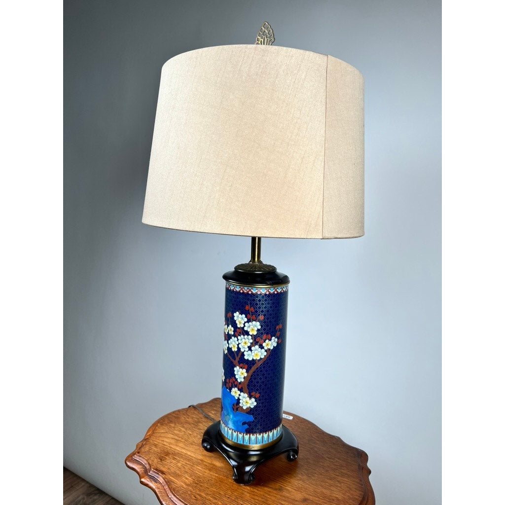 The Vintage Japanese Cloisonné Cylinder Table Lamp by Knob Creek, with a beige shade and blue floral cylindrical base, sits on a wooden side table against a plain gray wall.
