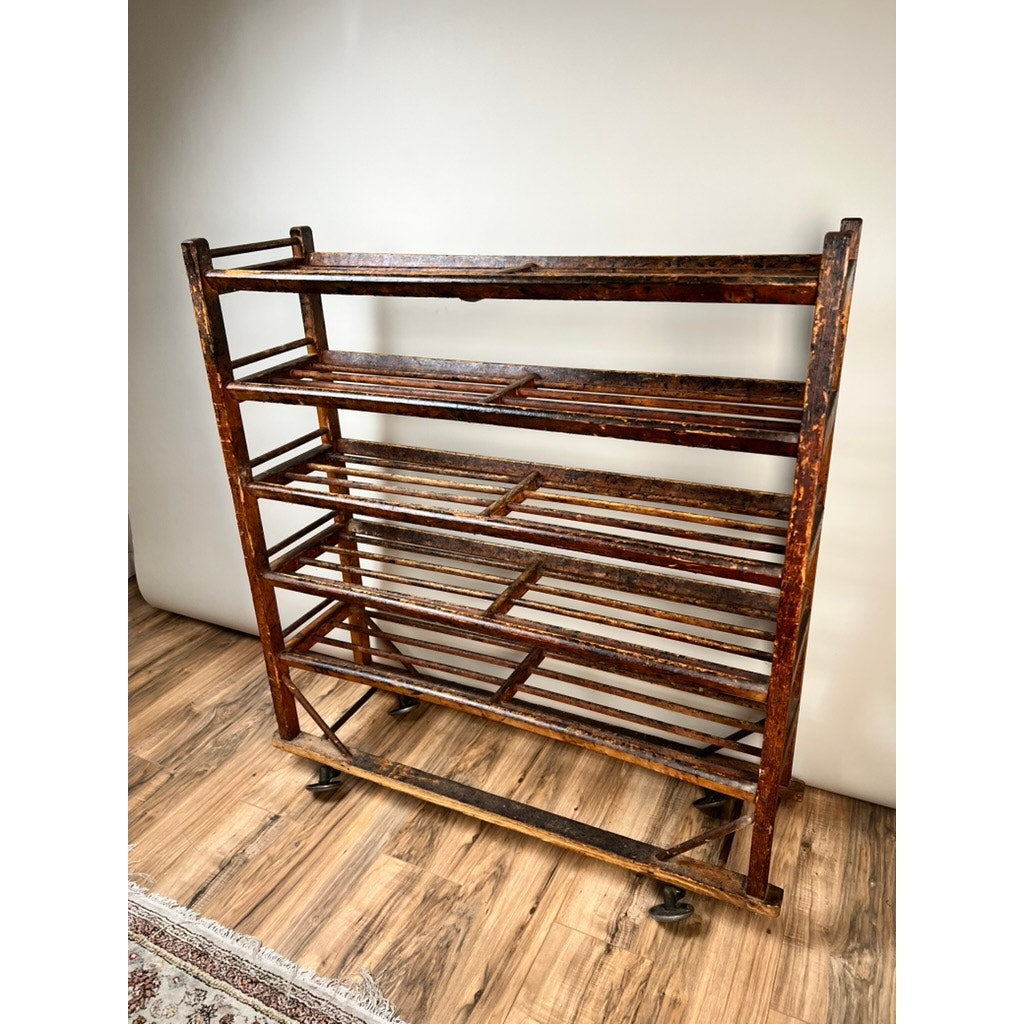 An Antique Oak Shoe Factory Rolling Rack on original industrial casters (late 19th century) with slatted shelves and a worn dark brown finish stands on a wood floor against a plain light-colored wall.
