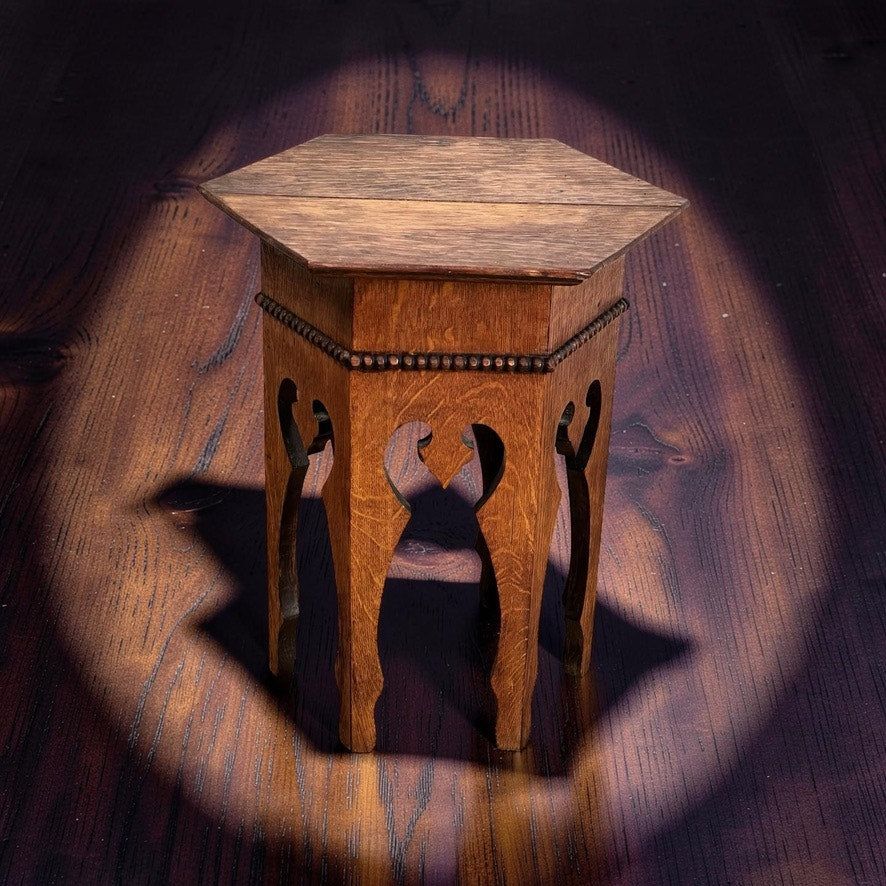 An Antique Arts & Crafts Small Oak Occasional Table c. 1910, featuring a hexagonal top and ornate cut-out legs, sits under a spotlight on a dark wooden floor, echoing classic Arts and Crafts furniture design.