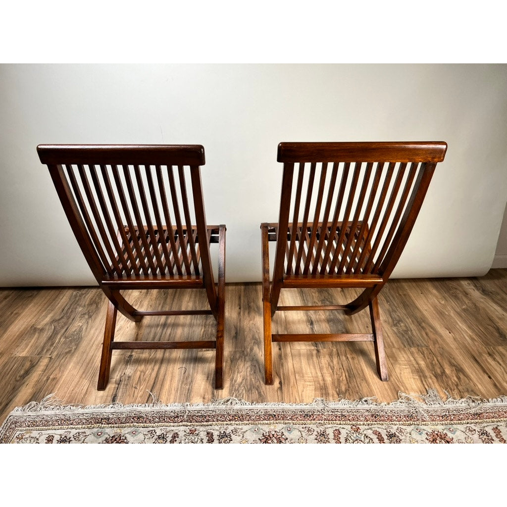 A Pair of Vintage Mid-Century Modern Style Teak Folding Chairs sits side by side on a wooden floor, with a patterned rug partially visible below and a plain light-colored wall in the background. Brand: None/Unknown.