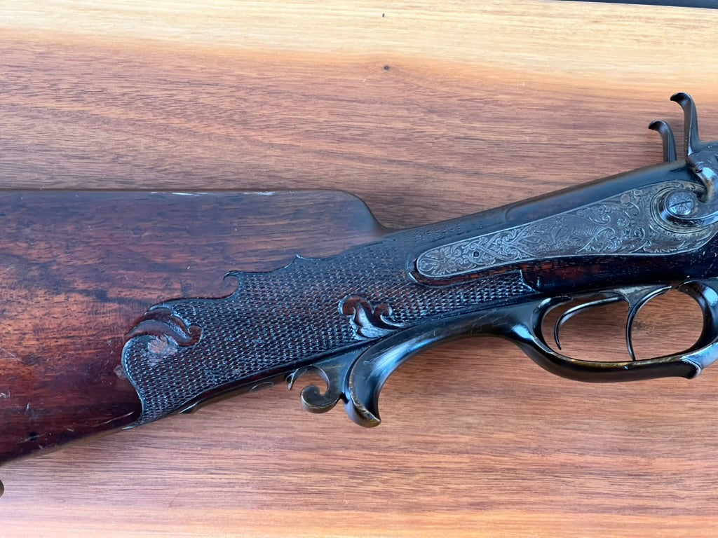 Close-up of the Antique Christoph Funke Suhl German Damascus Double Barrel Hammer Shotgun, c. 1880, from Pearl City Antiques, featuring ornate engravings on the metal and a dark carved wooden stock with intricate Damascus barrels, displayed on a light wood surface.