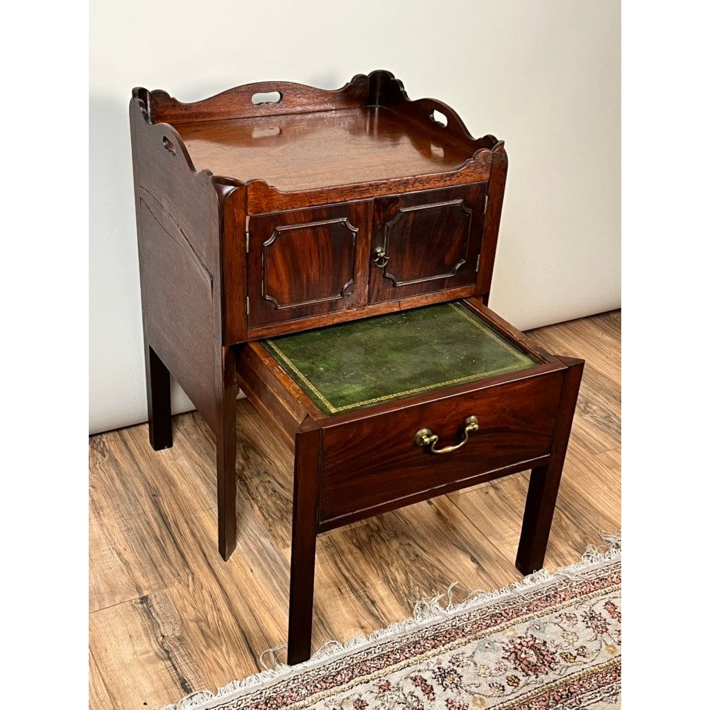 An Antique George III Bedside Commode c. Late 18th Century features ornate edges, two cabinet doors, and a drawer with a brass handle revealing a pull-out leather writing surface, standing on a wooden floor by a patterned rug.