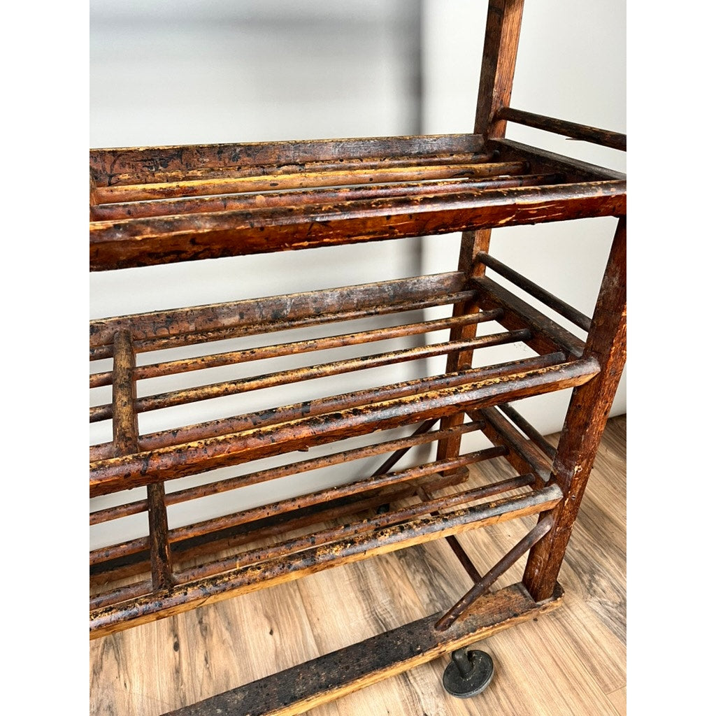 A close-up of an Antique Oak Shoe Factory Rolling Rack on original industrial casters (late 19th century), featuring three slatted shelves, visible scratches, chipped paint, and heavy use. Shown on a light wood floor by a white wall.