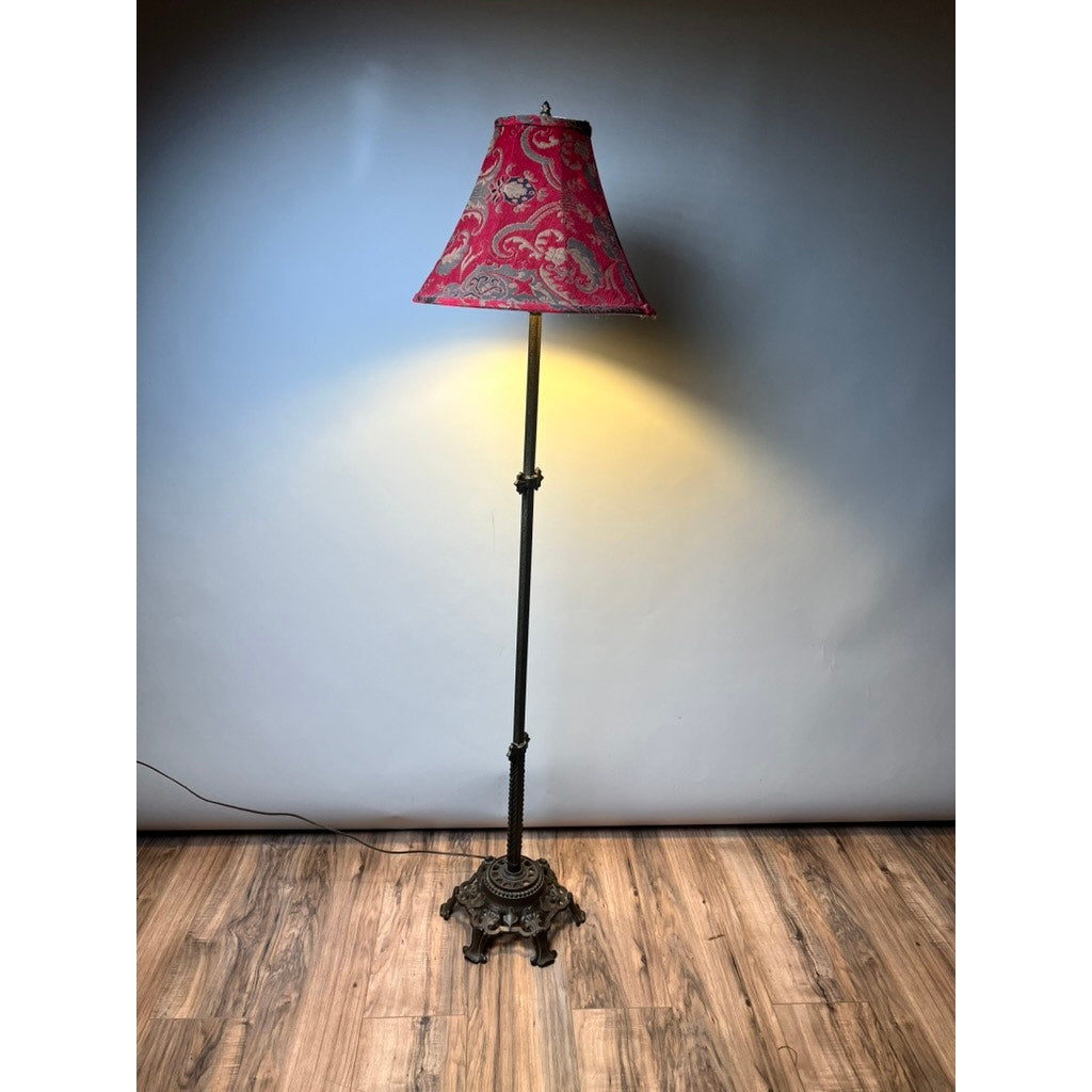 The Rembrandt Antique Art Deco Cast Iron Floor Lamp features an ornate base and a red shade, illuminating a wooden floor with warm light against a plain wall.