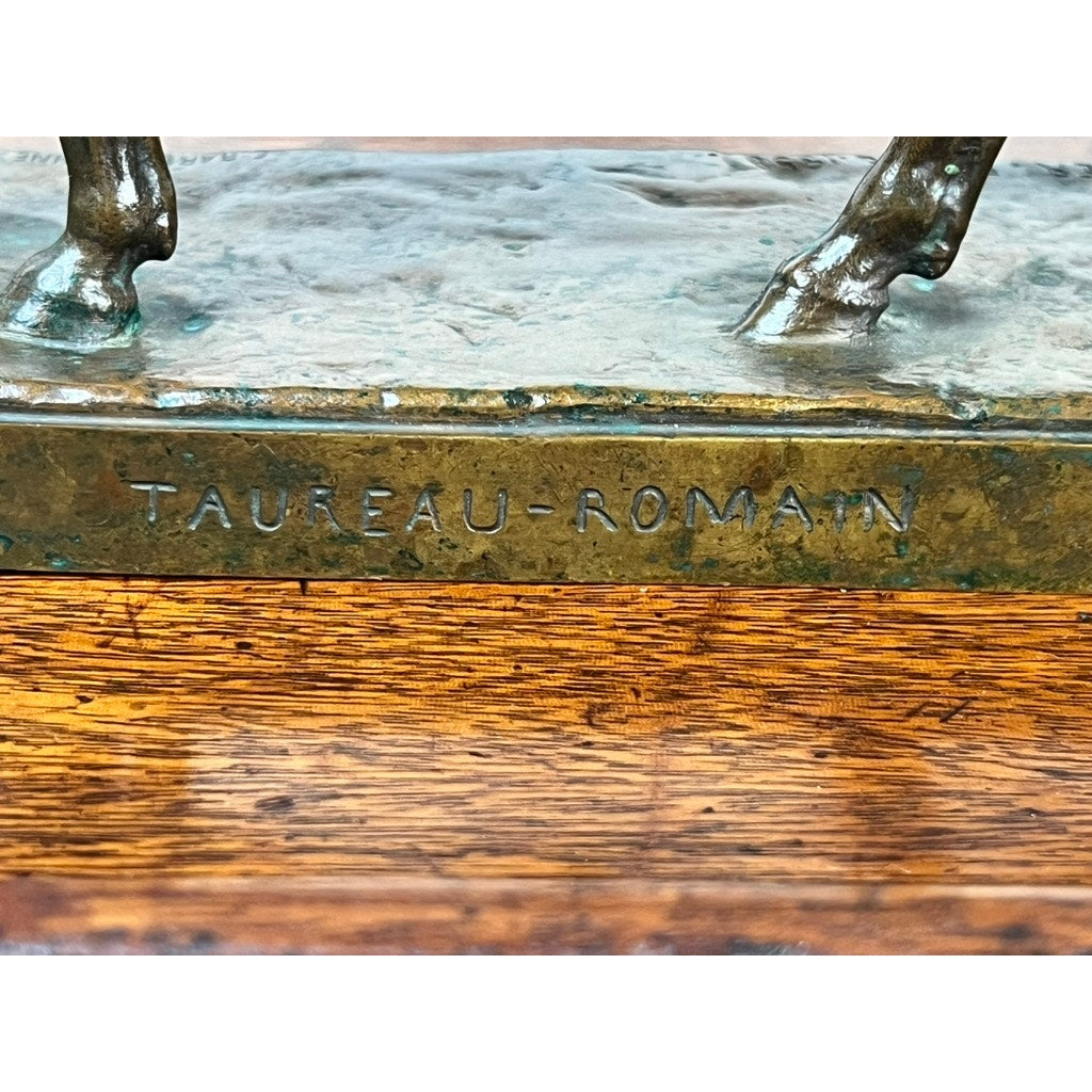 Close-up of the base of a Jean-Baptiste Clesinger “Taureau Romain” bronze sculpture (Barbedienne Foundry, Rome 1858), engraved with TAUREAU-ROMAIN and showing part of the animal’s lower legs, on a wooden surface.
