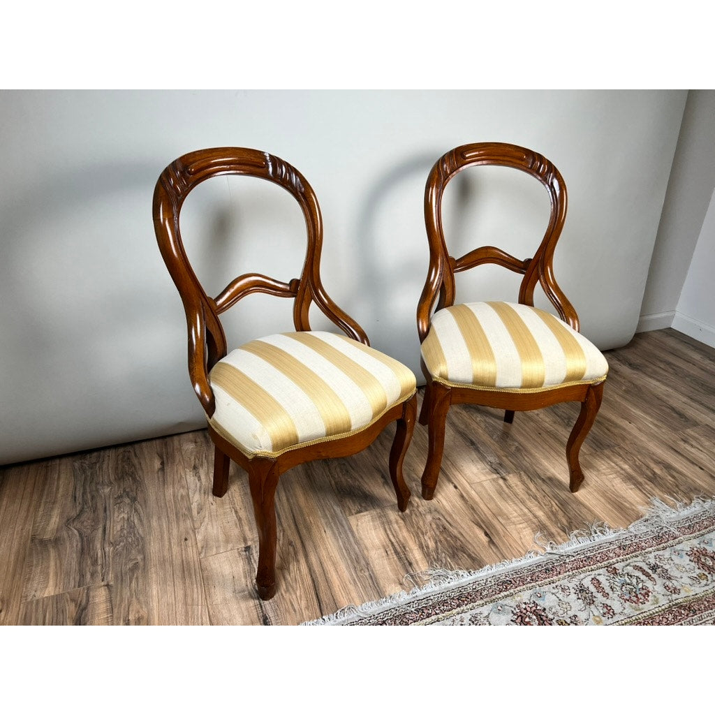 A pair of antique Victorian balloon back chairs (late 19th century) with yellow-and-white striped seats are placed side by side on a wooden floor beside part of a patterned rug and a plain white wall.