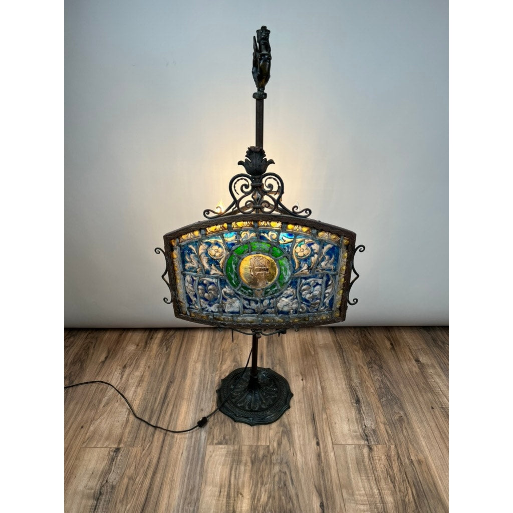 An Antique Five Burner Lucerne Whale Oil Lamp with a Napoleon I Fireplace Screen Shade, featuring ornate metalwork and stained glass in blue, green, and gold hues, stands on a wooden floor against a plain light-colored wall.