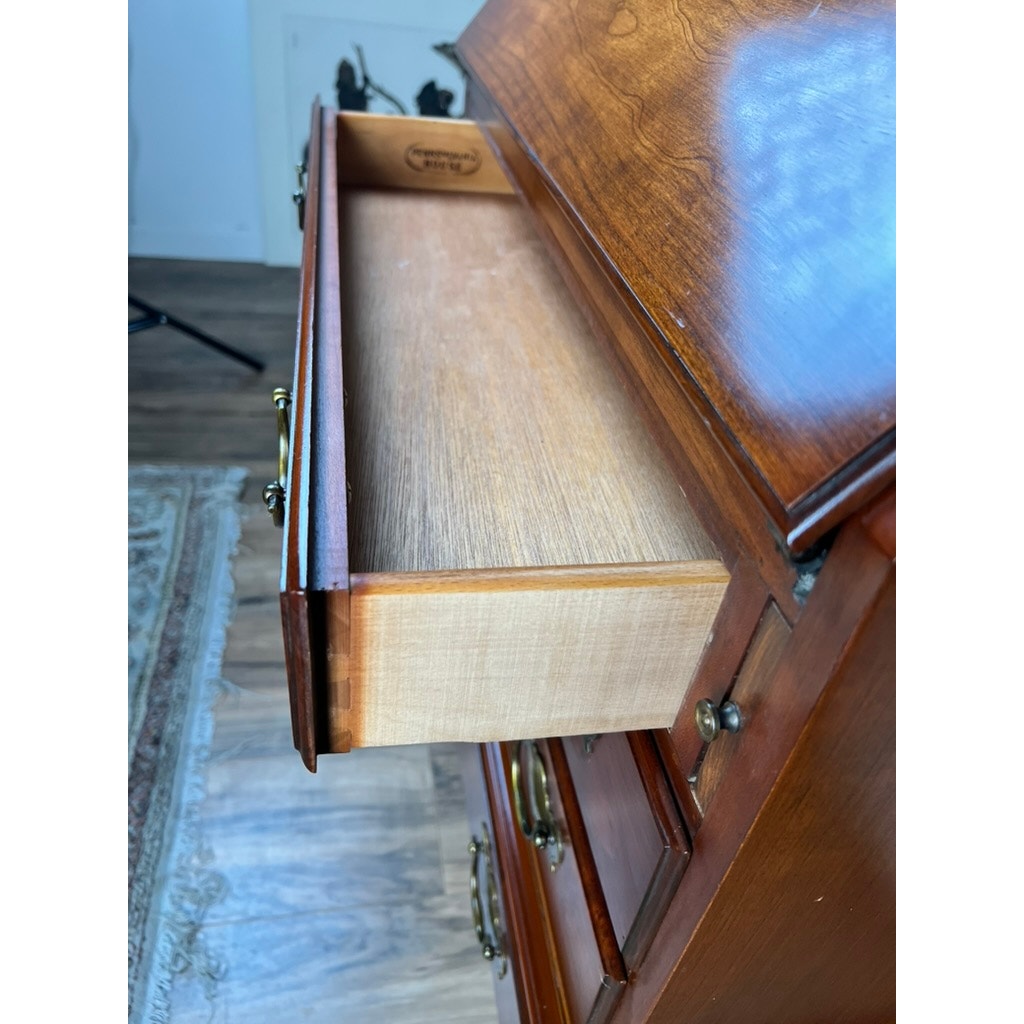 A close-up of a Vintage Solid Cherry Queen Anne Style Slant Front Secretary Desk by Pennsylvania House with an open drawer, revealing its light wood interior, brass handles, and glimpses of a rug and wooden floor in the background.