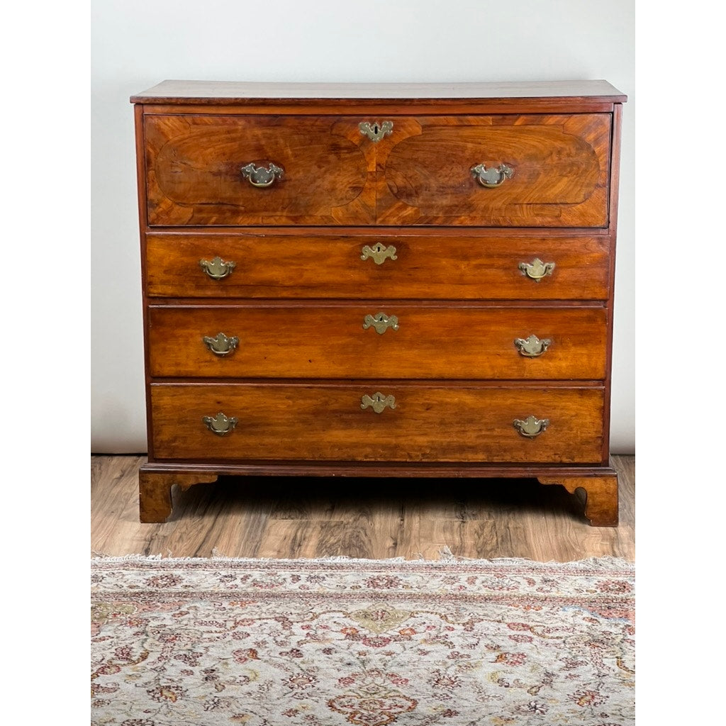 An Antique George IV Butlers Secretary from the early 19th century, featuring four drawers with brass handles, stands on a wood floor. Its rich brown finish contrasts a white wall, with part of a patterned rug visible below.