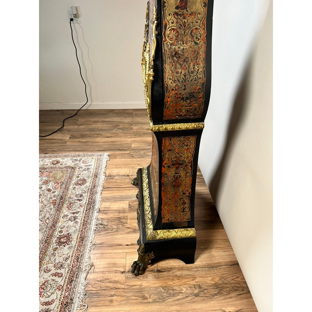 A Monumental French Napoleon III Boulle Marquetry Floor Clock with Ormolu Mounts, circa 1865–1885, featuring intricate gold and red marquetry, stands on a wood floor beside a patterned rug and a white wall with a visible electrical outlet and black power cord.