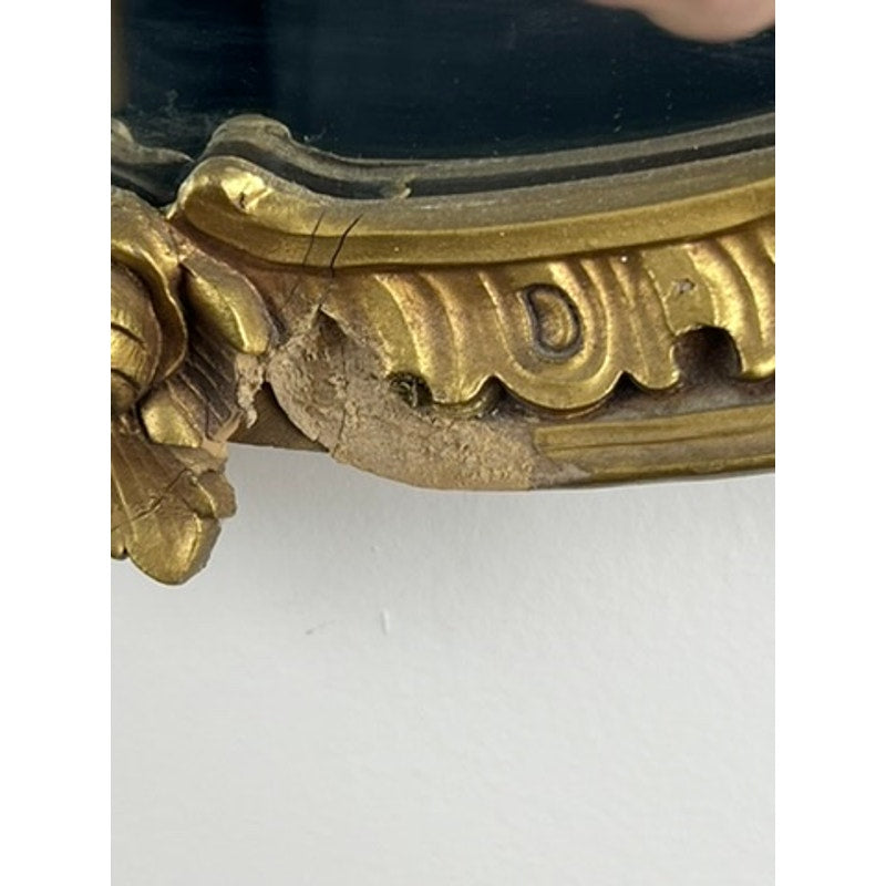 Close-up of an Antique Gilt Wood Oval Mirror (c. Early 20th Century) showing ornate classical revival details and visible chipping on the lower edge, exposing worn wood beneath the gilt finish. Brand: Unknown.