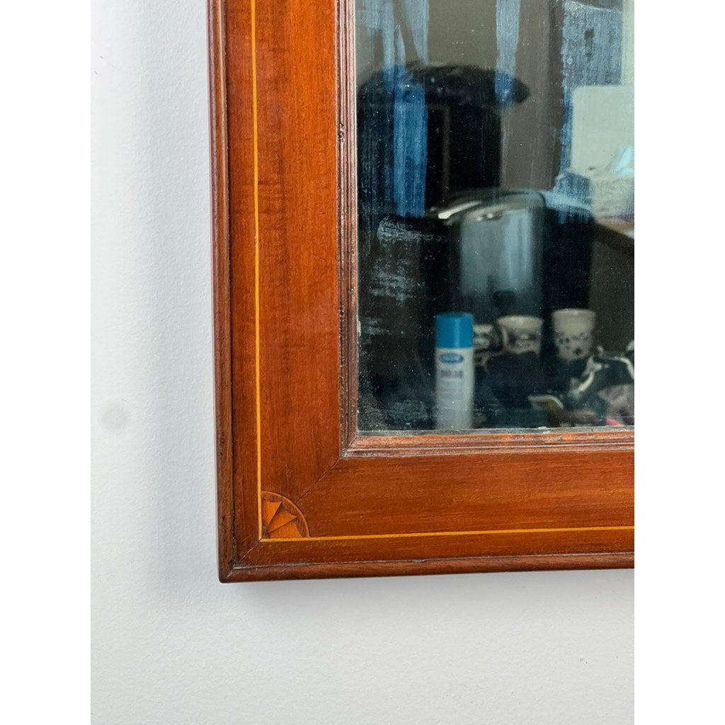 A close-up of the bottom left corner of an American Federal Mahogany Wall Mirror with Fan Inlay and original hand silvered glass, circa 1810–1825, shows its wooden frame reflecting various household items—including a can, cups, and a basket—against a white wall.