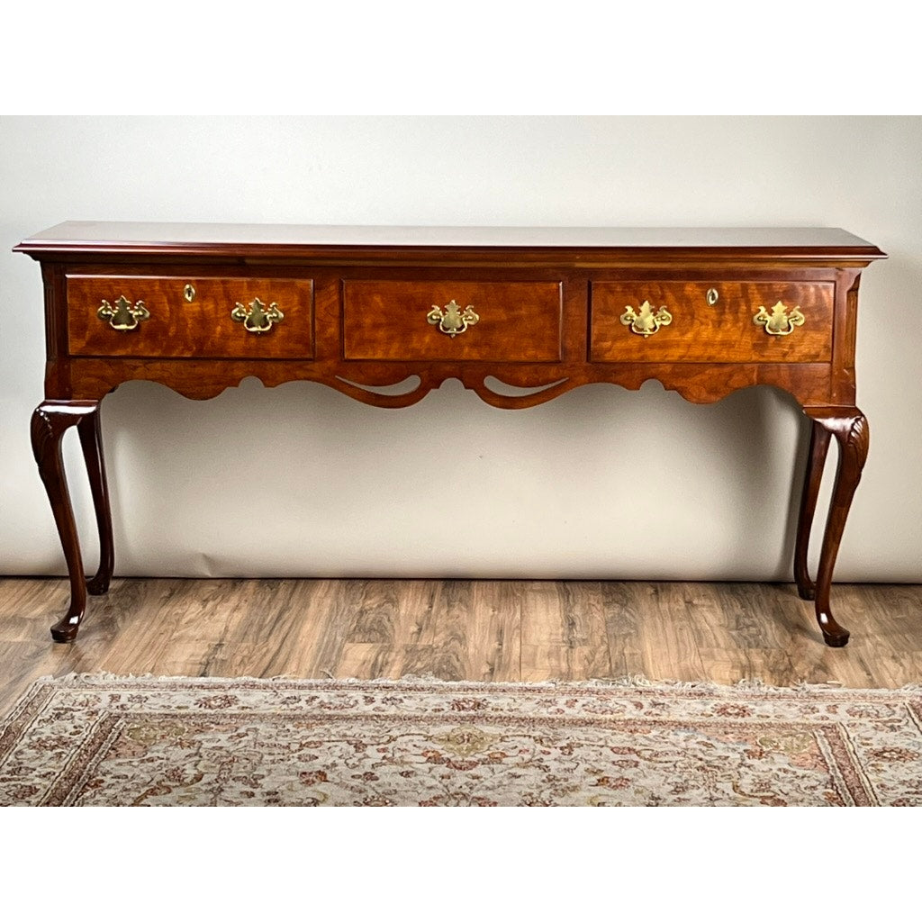 The Vintage Solid Cherry Queen Anne Style Sideboard by Henredon features four drawers with ornate brass handles, curved legs, and a polished finish, sitting on a wooden floor with part of a patterned rug in front.