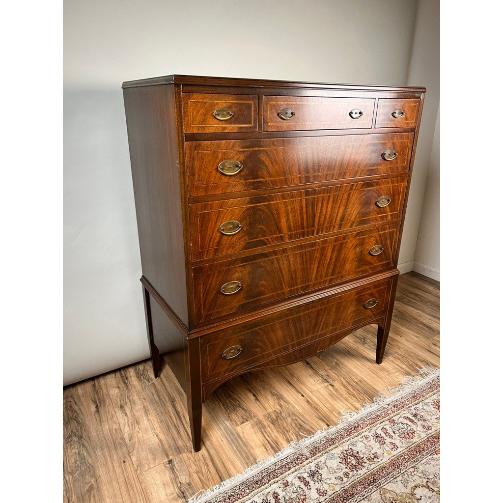 A Vintage Dutch Woodcraft Shops Federal Style Flame Mahogany Chest on Stand (c. 1945–1955) by Dutch Woodcraft Shops, featuring brass handles, stands on a wooden floor beside a patterned rug and is set against a plain white wall.