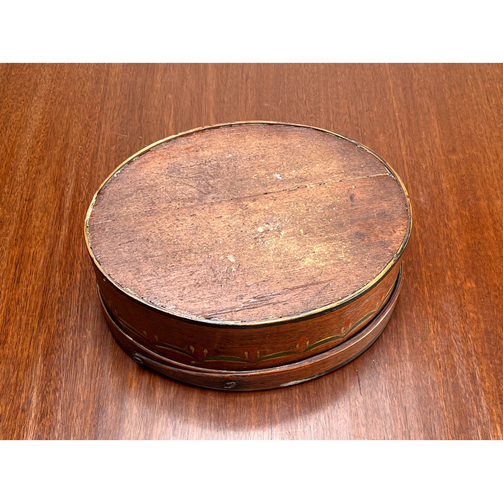An Antique German Polychrome Painted Bentwood "Spanschachtel" or Ribbon Box, c. late 18th century, with an oval form and worn flat lid, sits on a polished wooden surface. Fading and small marks on the lid highlight its rustic charm and age.