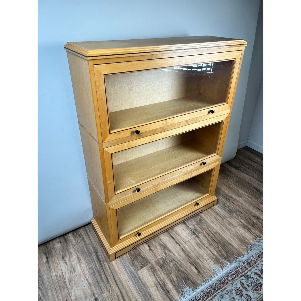 A Vintage Bench Made Maple with Wenge Inlay Three Stack Barrister Bookcase stands on a wood floor beside a white wall and the corner of a patterned rug, featuring glass-front sections with black knobs.