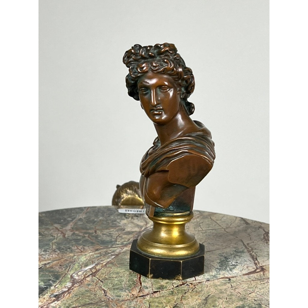 An antique small bronze bust of “the Apollo Belvedere,” marked by a Vienna foundry, sits on a round gold and black pedestal atop a marble surface against a plain background. Brand unknown.