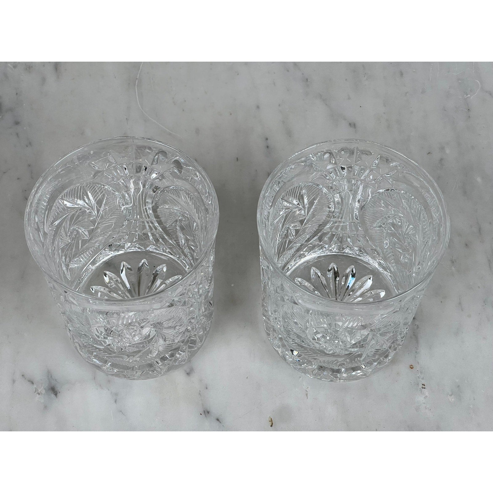 A pair of Fostoria Stowe Double Old Fashioned Tumbler Glasses with decorative patterns sit side by side on a light gray marble surface, evoking vintage American glassware.