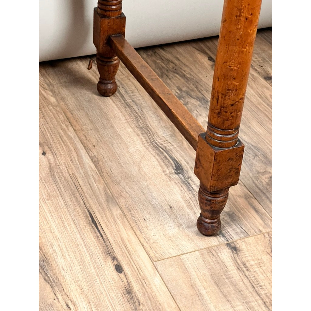A close-up of the leg of an Antique Sheraton Birdseye Maple One Drawer Stand (early 19th c.), featuring carved details and a connecting horizontal wooden support, standing on a light brown wood floor. Brand: unknown.