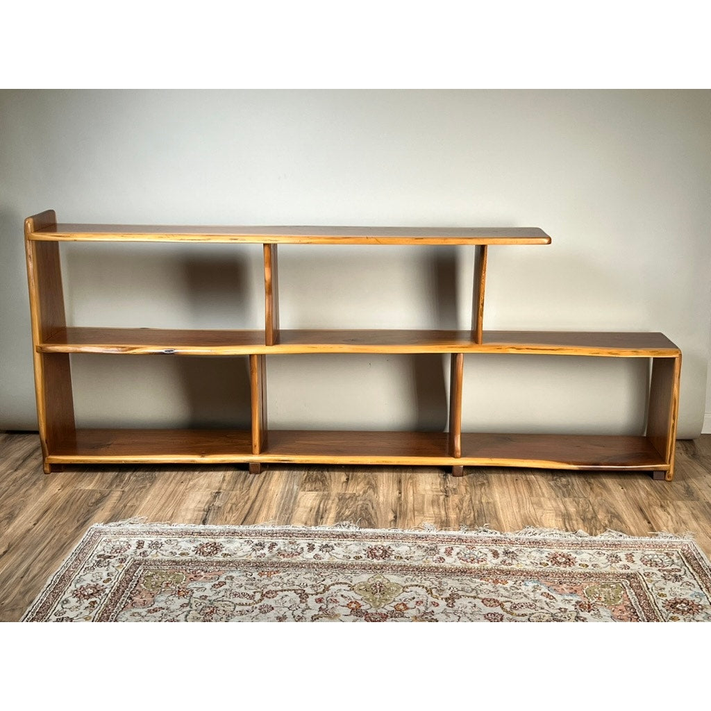 A Vintage Mid-Century Modern Style Live Edge Walnut Bookshelf with an asymmetrical three-tier design and open compartments stands on a wood floor with a patterned rug and a plain light-colored wall behind. Brand: None or Unknown.