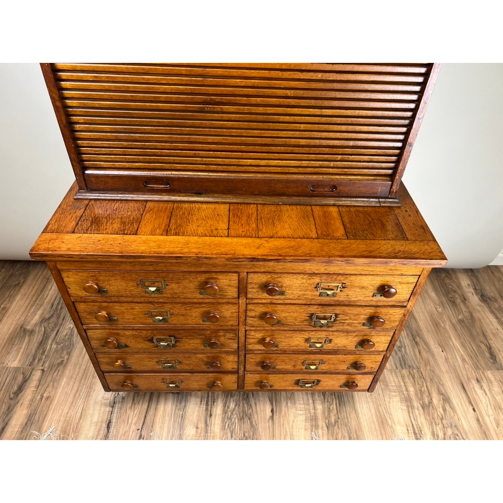 An Antique Early 20th Century Oak Multidrawer File with Roll Top Pigeon Hole Top stands on a wood floor, its small drawers fitted with round wooden knobs and brass label holders, capturing the charm of an antique office organizer. Brand: None/Unknown.