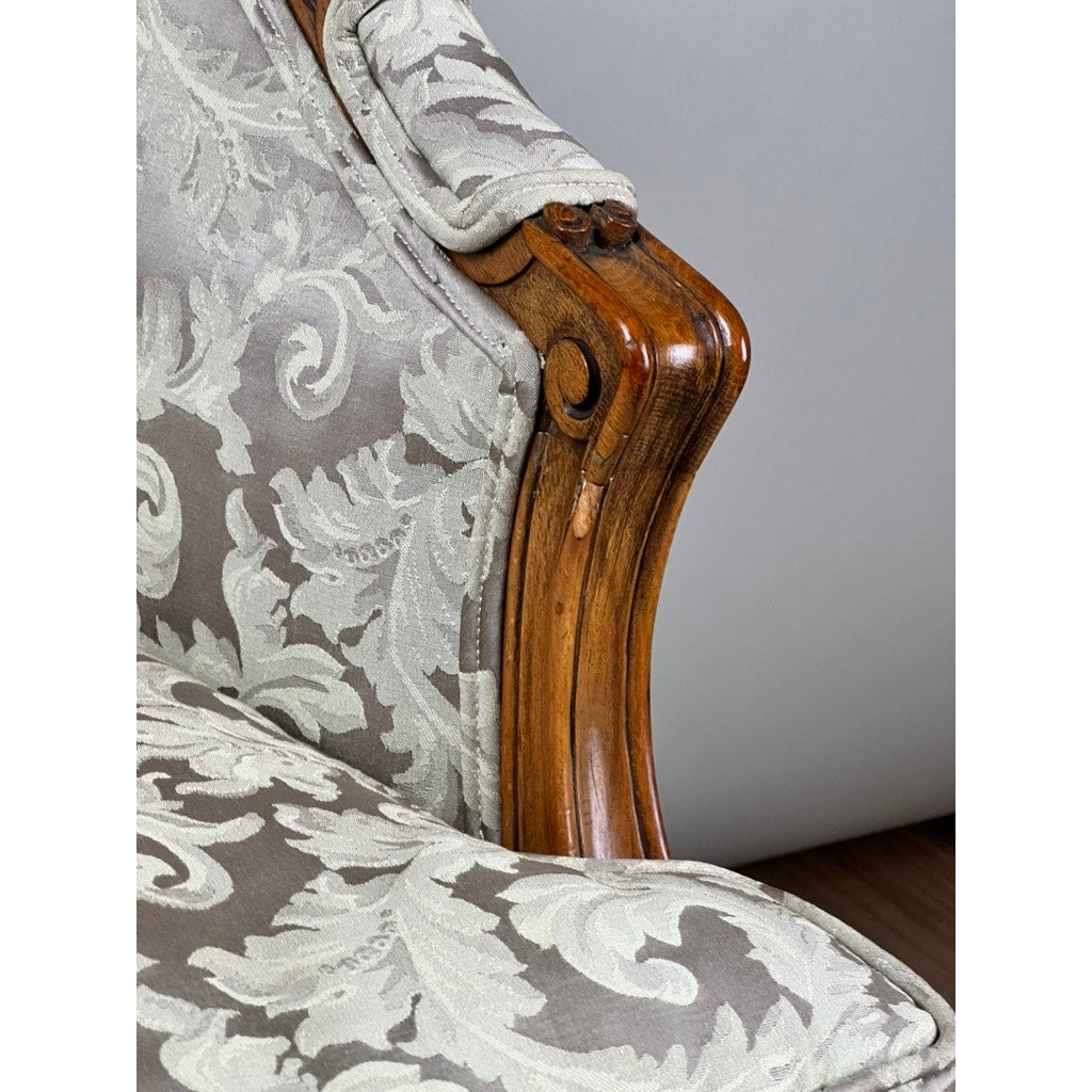 Close-up of a carved walnut armrest on an Antique French Louis XV Rococo Style Bergère Arm Chair (mid 19th c.), featuring polished, slightly worn wood and newer light grey floral damask upholstery. Brand: Unknown.