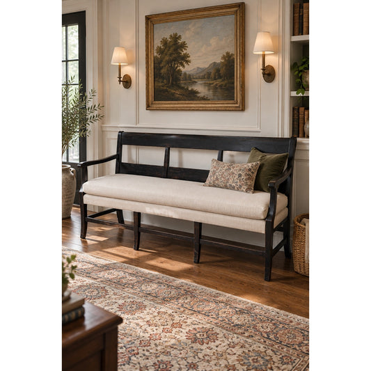 The Pearl City Antiques “Antique English Oak Three-Seat Bench, Late 19th Century,” featuring a beige cushion and pillows, sits against a wall adorned with a landscape painting and two wall sconces, in a cozy room with a patterned rug and bookshelves.