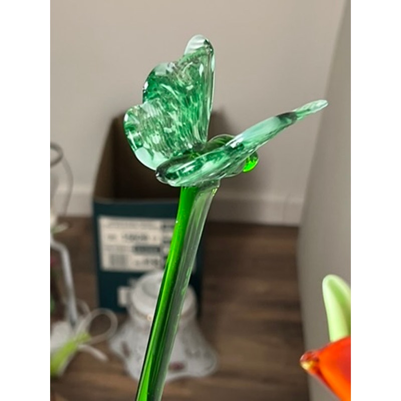 A close-up of a green glass butterfly perched on a thin stem, part of the Vintage Bouquet Murano Art Glass Long Stem Flowers and Butterflies, with a blurred background showing a box and other objects.