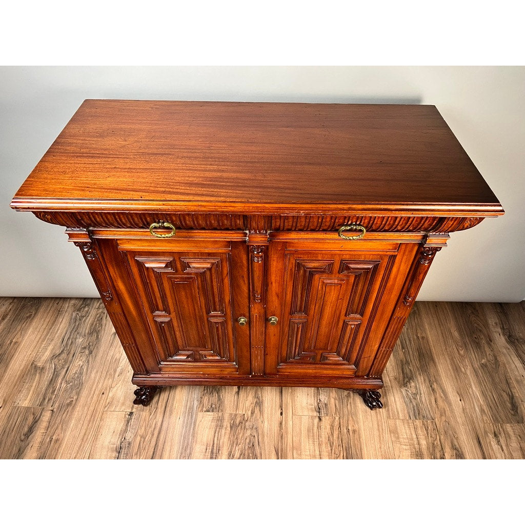 An Antique English Jacobean Revival Mahogany Buffet (c. 1915–1925) with ornate carvings, two cabinet doors, brass handles, and claw-style feet stands on a wood-patterned floor against a plain light wall.