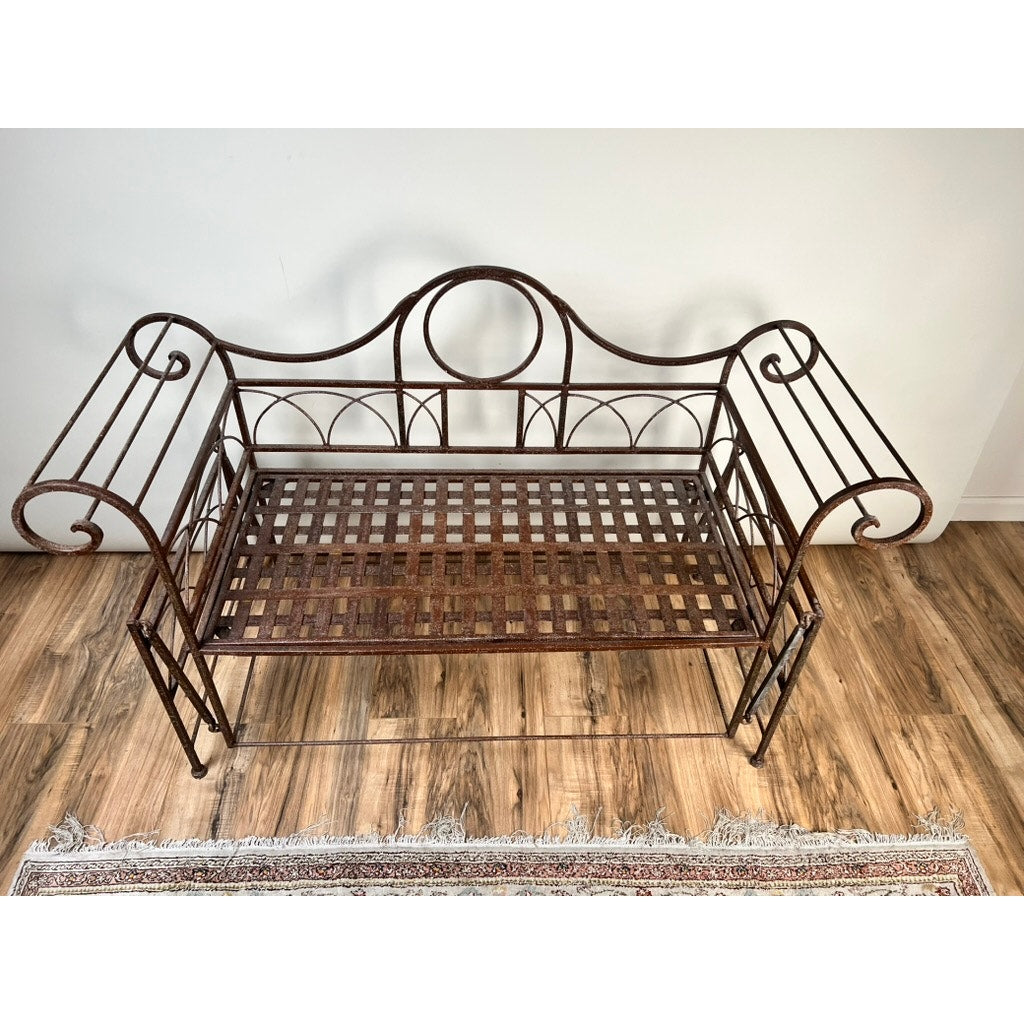 The Vintage Regency Style Wrought Iron Porch Glider, with ornate curved armrests and a lattice seat, sits on a wooden floor beside a fringed rug, adding vintage charm to the garden space against a plain white wall.