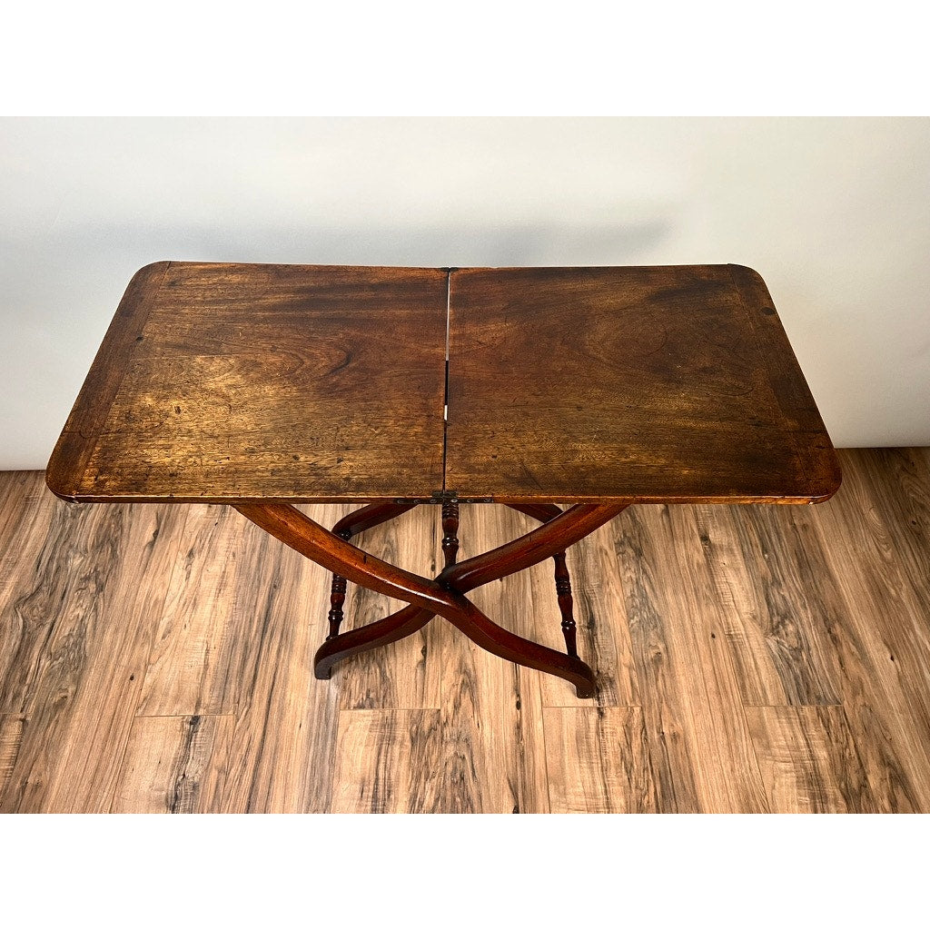 An Antique English Regency Mahogany Folding Coaching or Sporting Table (c. 1825–1845) with a rectangular top and X-shaped support stands elegantly on a wooden floor against a light-colored wall. Brand is unknown.