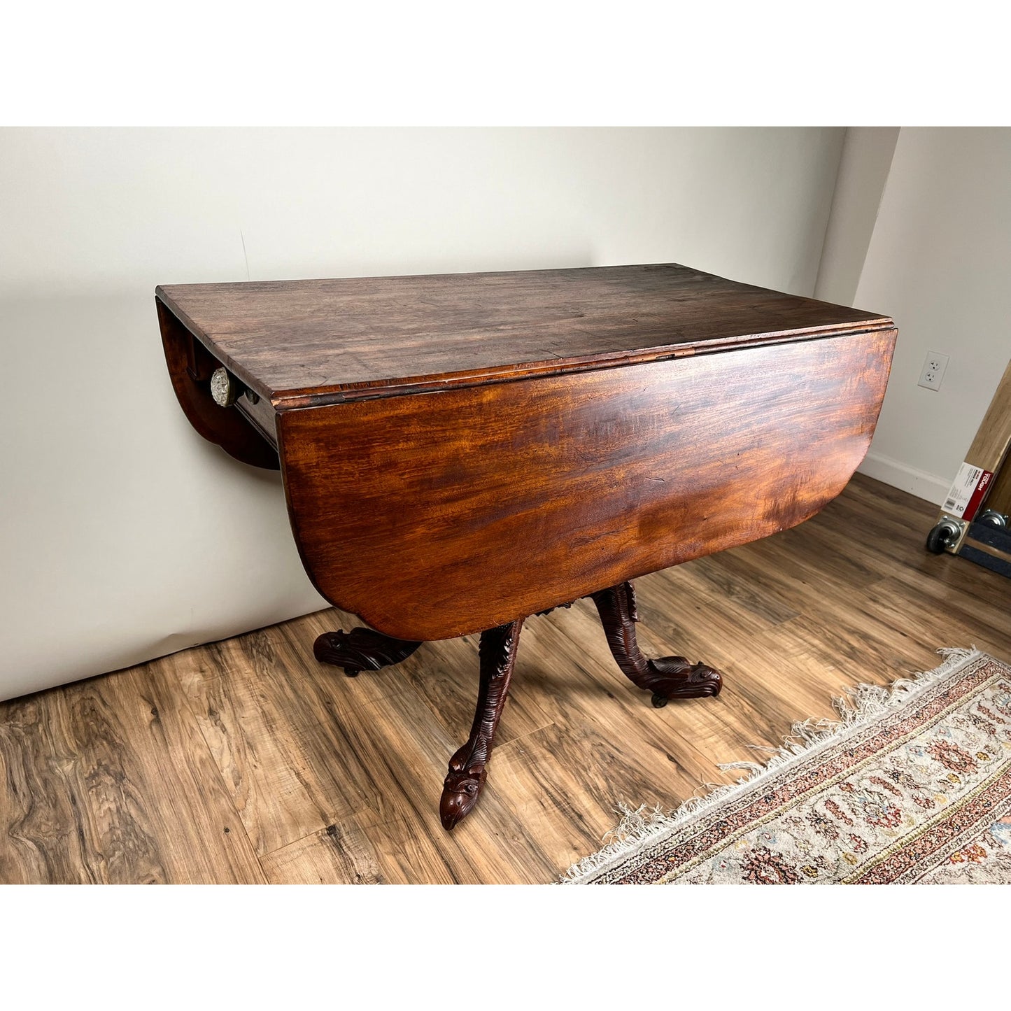 An Antique American Empire Drop Leaf Table with Great Eagle Carved Feet c. 1840 stands on a wood floor near a wall and a patterned rug, showcasing its dark finish and elegant curved sides.