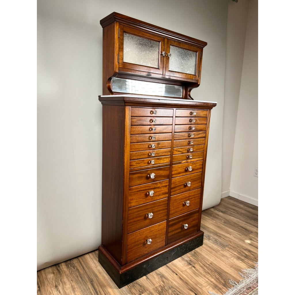 The Antique Mahogany Multi-drawer Dental Cabinet by The American Cabinet Co. (early 20th c.) features a black base, mirrored backsplash, two glass-panel doors, and multiple drawers, and stands on a wood floor against a white wall.