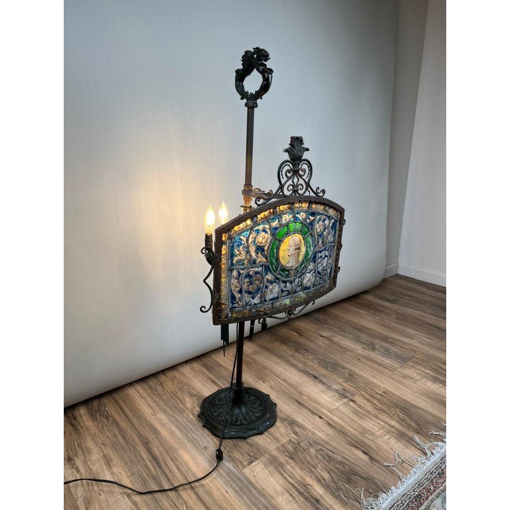 An Antique Five Burner Lucerne Whale Oil Lamp with a Napoleon I Fireplace Screen Shade stands on a wooden floor near a white wall, its ornate metal frame and colorful panels evoking Napoleonic elegance.