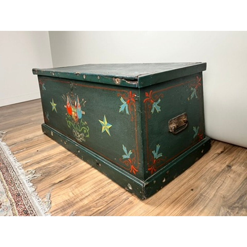 An Antique Paint Decorated English Sea Chest from the late 19th century, featuring hand-painted floral designs and stars in red, yellow, and blue on green wood, sits on a hardwood floor beside a patterned rug. Brand: Unknown.