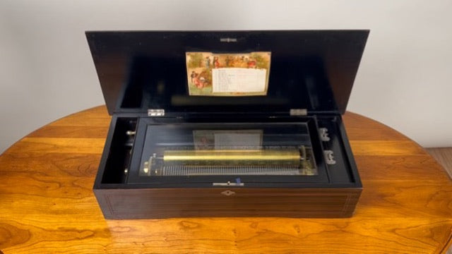 An Antique Swiss 12 Tune Music Box from the late 19th century sits open on a polished wooden surface, revealing its metal cylinder and comb mechanism, with a decorative card attached inside the lid. Brand unknown.