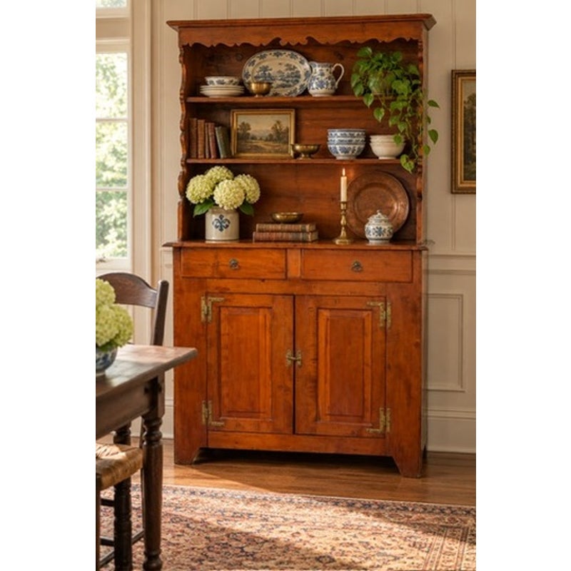 A tall 19th-century Pennsylvania or Mid-Atlantic Cherry Open Pewter Cupboard with open shelving at the top displaying decorative plates, books, and plants, over a two-drawer, two-door cabinet base.