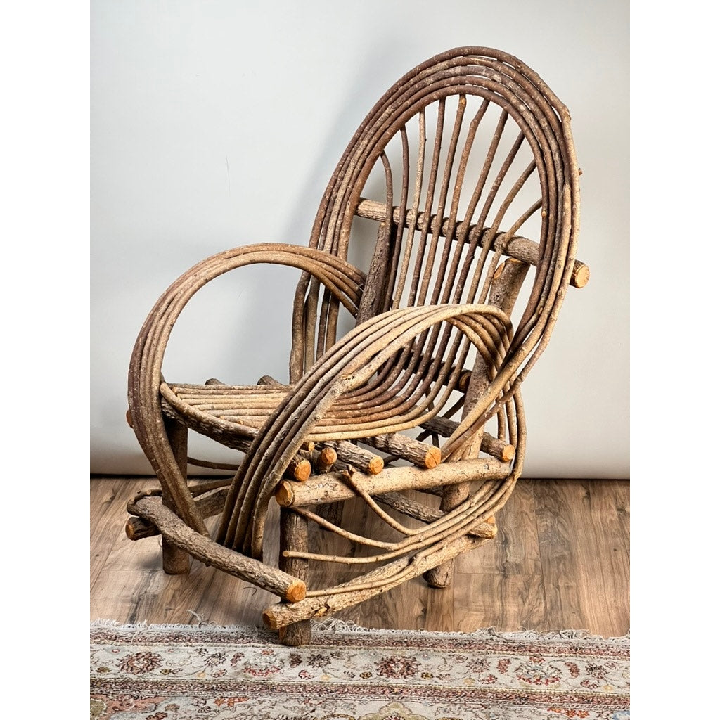 The Vintage Adirondack Bent Willow Arm Chair, crafted from curved, unfinished wood branches, rests on a wooden floor with an ornate rug partially visible in front and a plain light background. Brand: None or Unknown.
