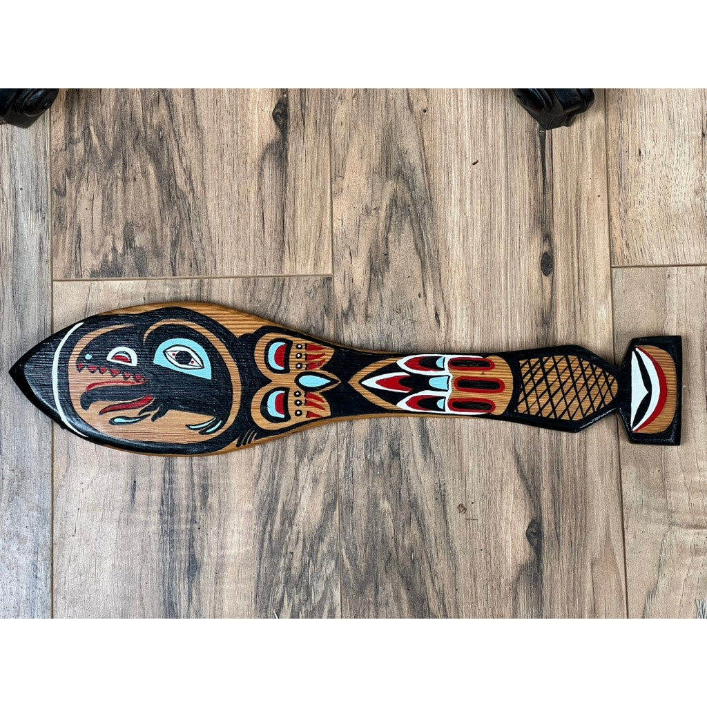 A Vintage Inuit Carved Red Cedar Paddle from Sitka, Alaska, signed illegibly, featuring stylized animal art and bold black, red, and white patterns, rests on a wood-grain floor. Brand unknown.