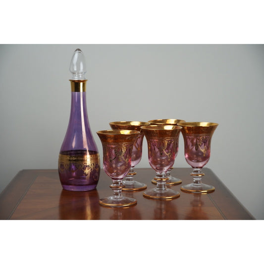 A Vintage Six Piece Italian Amethyst Glass Decanter Set with 22k Gold Accents by Pearl City Antiques, c. 1960, is displayed elegantly on a polished wooden table. The striking purple and gold decanter with stopper is accompanied by five matching goblets, all set against a plain light background.