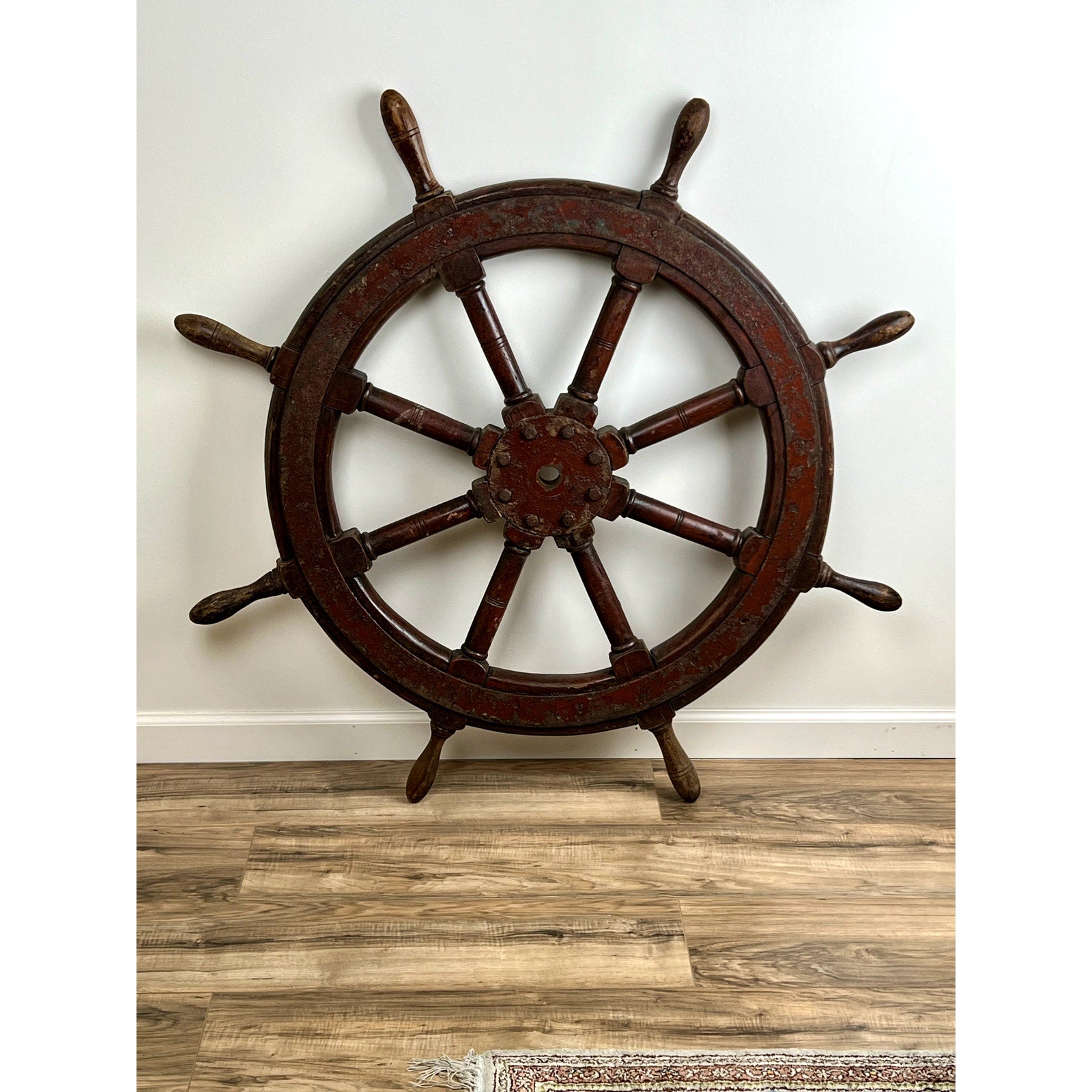 An Antique Eight Spoke Mahogany and Iron Ship’s Wheel stamped “1864 US” rests against a white wall on a wooden floor, part of a rug visible in the corner, evoking classic 19th-century maritime style.